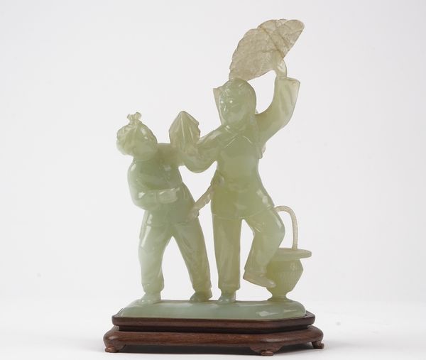 A Chinese bowenite group of a boy and girl, 20th century, carved standing, the girl holding aloft a fan, a basket at her feet, 23cm. high, wood stand,