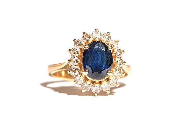 A gold, sapphire and diamond oval cluster ring, claw set with the oval cut sapphire in a surround of circular cut diamonds, detailed 14 K, ring size M