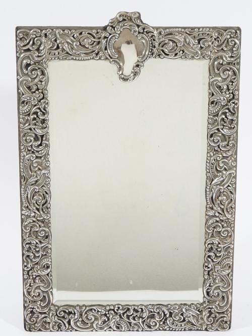 A silver mounted shaped rectangular strut backed mirror, fitted with a bevelled mirror glass, the wide border embossed and pierced with foliate scroll