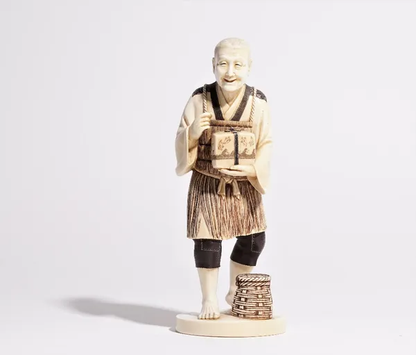 A Japanese ivory okimono of a man, carved standing holding a box, a basket at his feet, 21cm high.