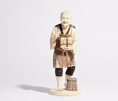 A Japanese ivory okimono of a man, carved standing holding a box, a basket at his feet, 21cm high.