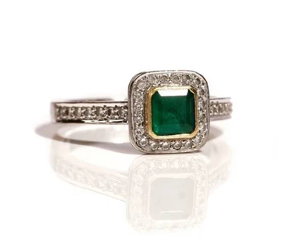 An 18ct white gold, emerald and diamond cluster ring, mounted with the cut cornered square step cut emerald to the centre, in a surround of circular c