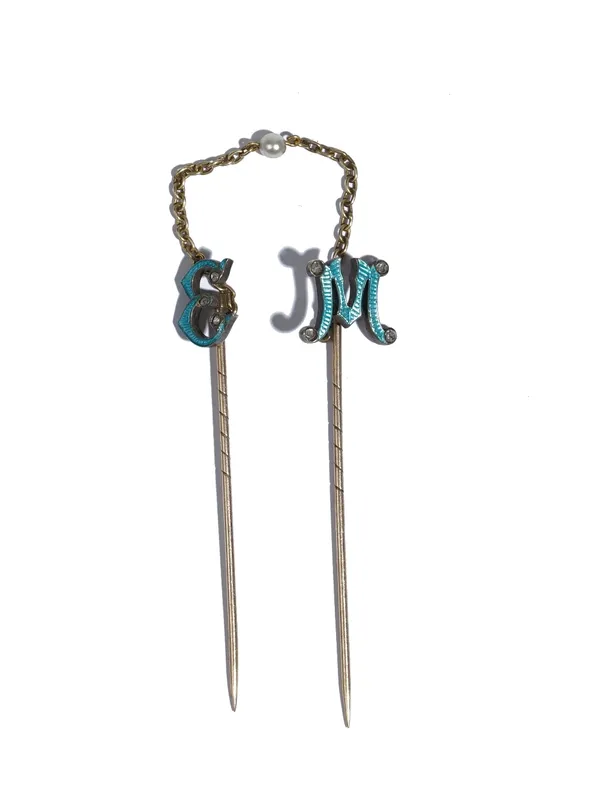 Two diamond set and pale blue enamelled stick pins, one designed as the initial 'E', the other designed as the initial 'M', the connecting chain mount