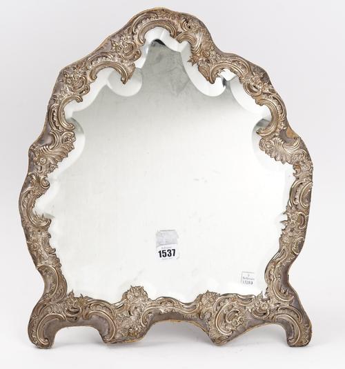A late Victorian silver mounted strut backed mirror, of shaped rococo form, the mount with floral and scroll pierced and embossed decoration, fitted...