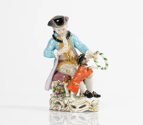 A Meissen figure of a seated boy, late 19th century, holding out a striped hoop through which his dog, dressed in a tricorn hat and red jacket, is...
