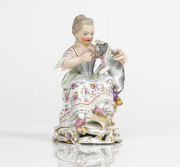 A Meissen figure of a seated girl, late 19th century, modelled holding a grey cat wearing a cap and feeding it with a spoon, blue crossed swords...