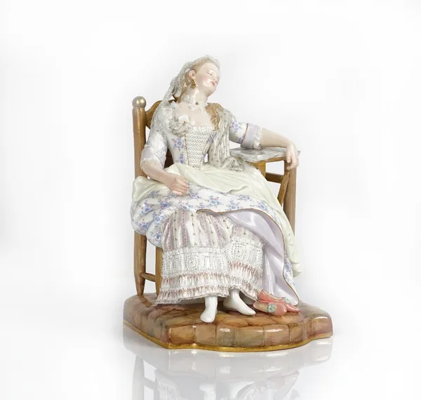 A Meissen figure of a lady, late 19th century, seated asleep in a chair, her left arm resting on a table, a single playing card slipped inside her...