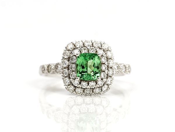 A white gold, tsavorite garnet and diamond cluster ring, claw set with the cushion shaped tsavorite garnet in a two tier surround set with circular...