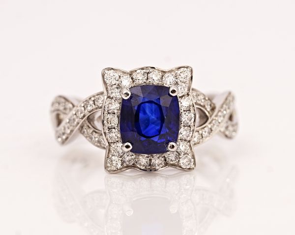 A platinum, sapphire and diamond cluster ring, claw set with the cushion shaped sapphire in a shaped rectangular surround set with circular cut...