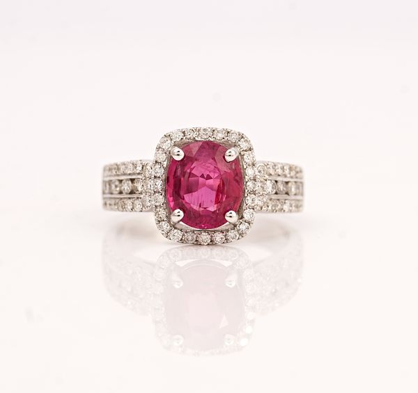 A white gold, ruby and diamond ring, claw set with the oval mixed cut ruby in a surround of circular cut diamonds, between diamond set shoulders in a...