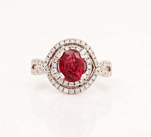 An 18ct white gold, ruby and diamond ring, claw set with the oval cut ruby in a surround of circular cut diamonds and with an outer border of...