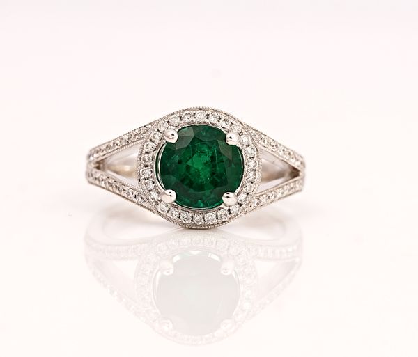 An 18ct white gold, emerald and diamond cluster ring, claw set with the circular cut emerald in a halo surround set with circular cut diamonds,...
