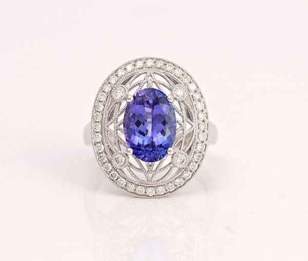 A white gold, tanzanite and diamond ring, claw set with the oval cut tanzanite within a lattice work surround, in a border of circular cut diamonds,...