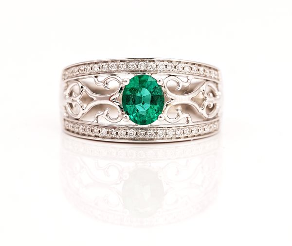 A white gold, emerald and diamond ring, claw set with the oval cut emerald between scroll pierced sides and between two rows of circular cut...