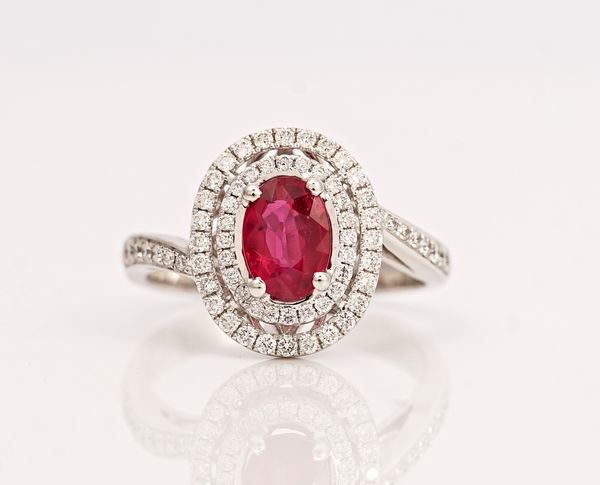 A white gold, ruby and diamond oval cluster ring, claw set with the oval cut ruby in a two tier oval surround, set with circular cut diamonds,...